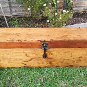 May include: A rectangular wooden chest with a hinged lid and a metal latch. The chest is made of light-coloured wood with a darker wood accent on the top. It sits on green grass.