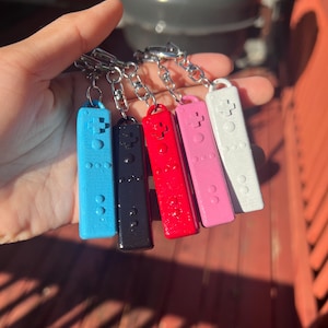 May include: A selection of six keyrings in the shape of game controllers. The keyrings are in blue, black, red, pink, and white. Each keyring has a silver chain and a ring for keys. The keyrings are made of a solid material.