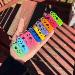 May include: A collection of colorful, miniature game controller earrings in various colors, including green, blue, pink, orange, yellow, and purple. Each controller has black button details and a silver hoop for the earring hook. The earrings are held in a hand.