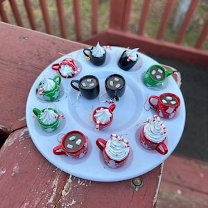 May include: A selection of handmade earrings resembling miniature mugs of hot chocolate, adorned with whipped cream and candy cane details. The earrings come in various colours, such as red, green, and black, and are presented on a white plate.