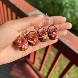 May include: Handmade earrings featuring miniature brown baskets filled with brown bread, lined with red and white checkered fabric. The earrings have silver-colored hooks and are held in a hand against a blurred background.