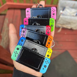 May include: A collection of colorful miniature game console keychains. Each keychain features a black console body with vibrant side controllers in pink, green, blue, purple, orange, and yellow. The keychains are held in a hand against a blurred outdoor background.