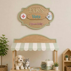 May include: A wooden veterinary play stand with a striped awning, featuring a sign that reads Evie's Vets Open daily. The set includes a plush dog, a plush cat, a toy pet carrier, and a toy stethoscope, all arranged on a wooden tabletop surface.