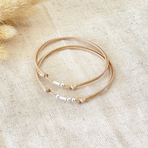 May include: Two beige string bracelets with silver beads. The bracelets are stacked on top of each other.