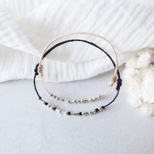 Könnte beinhalten: Zwei handgefertigte Armbänder. Eines ist schwarz, das andere beige. Beide haben silberne Perlen und sind mit Knoten gebunden. Die Armbänder liegen auf einer hellgrauen Oberfläche mit einem weißen Tuch und weißen Blumen im Hintergrund.