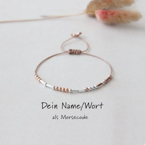 Könnte beinhalten: Ein zartes Armband mit braunem Band und silbernen und roségoldenen Perlen. Das Armband ist verstellbar und kann mit einem Namen oder Wort in Morsecode personalisiert werden. Der Text "Dein Name/Wort als Morsecode" steht unter dem Armband.