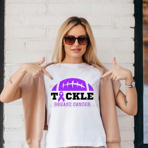 Op de afbeelding: Wit T-shirt met een paarse American football afbeelding en de tekst "TACKLE BREAST CANCER". Het ontwerp bevat een paars lint, een symbool van borstkankerbewustzijn. Een casual kledingstuk.