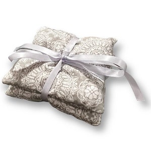 May include: Two gray and white patterned fabric squares tied together with a white satin ribbon. The squares are filled with a material that can be heated or cooled for therapeutic purposes.