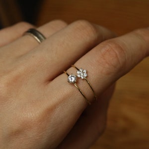 Puede incluir: Dos anillos de oro con pequeñas piedras preciosas transparentes. Un anillo tiene una sola piedra redonda, el otro tiene un grupo de piedras en forma de flor.