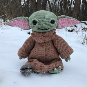 May include: A crocheted plush toy of a popular character from the Star Wars franchise. The toy is wearing a brown knitted sweater and has large green eyes and pink ears. The toy is sitting in the snow.