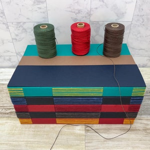 May include: A wooden box with a multi-colored striped design. Three spools of thread are on top of the box, one green, one red, and one brown.