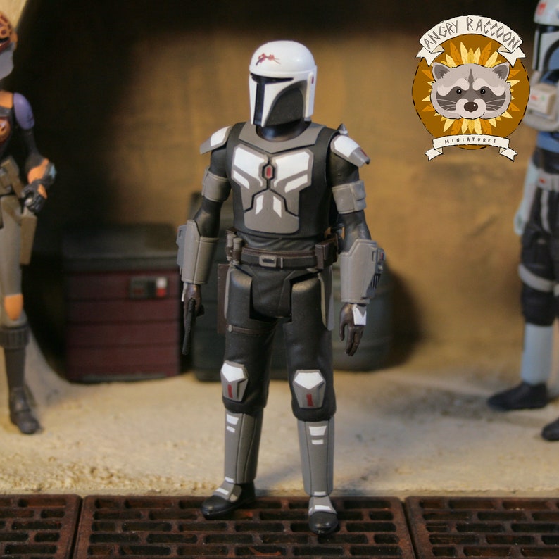 May include: A Mandalorian action figure in a black, gray, and white armored suit, holding a blaster pistol. The figure has a white helmet with a T-shaped visor. The Angry Raccoon Miniatures logo is in the background.