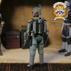 May include: A detailed action figure of a character from the Star Wars universe, viewed from the back. The figure is primarily green and grey, with orange accents on the helmet. It has a jetpack and various accessories. The Angry Raccoon Miniatures logo is visible.