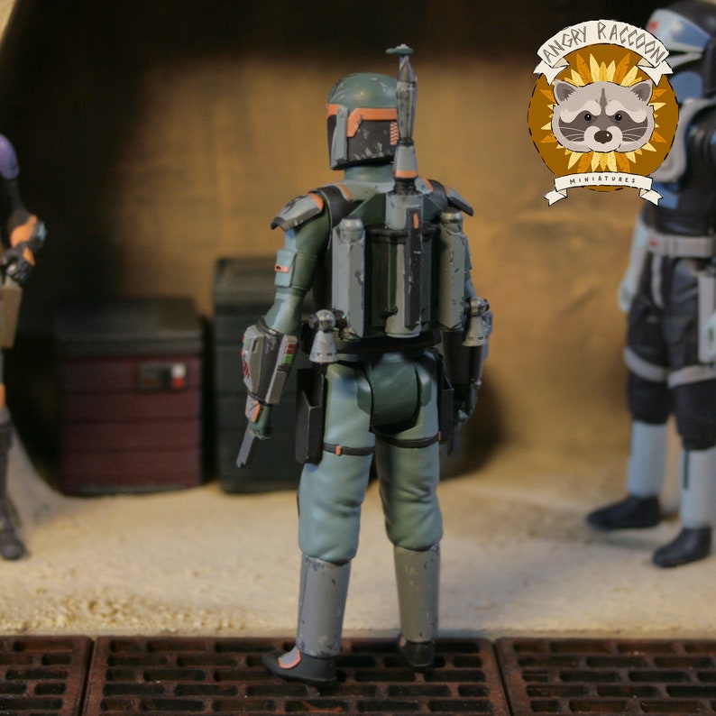 May include: A detailed action figure of a character from the Star Wars universe, viewed from the back. The figure is primarily green and grey, with orange accents on the helmet. It has a jetpack and various accessories. The Angry Raccoon Miniatures logo is visible.