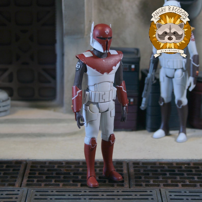 May include: A standing action figure of a soldier wearing white and red armour with a matching helmet. The figure features dark brown undersuit sleeves, red gauntlets, and red boots. A circular logo with a raccoon illustration and the text ANGRY RACCOON MINIATURES is visible in the upper right corner.