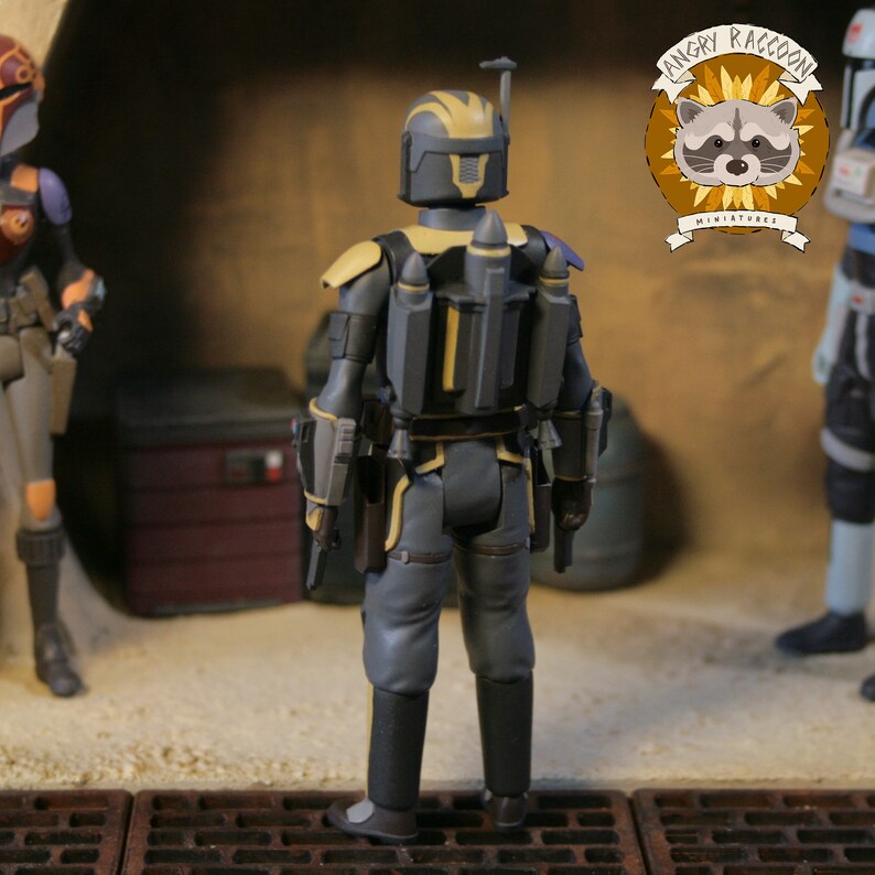 May include: A detailed action figure of a character in a gray and yellow armored suit, viewed from the back. The figure has a jetpack and holsters. The Angry Raccoon Miniatures logo is visible in the background.