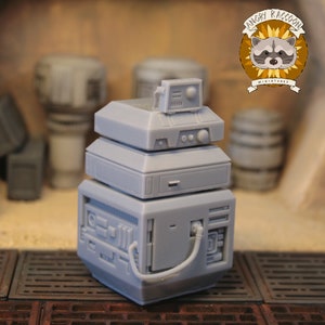 Peut inclure: Une maquette grise d'un appareil futuriste en couches. Le modèle a une forme carrée avec des panneaux détaillés, un petit écran et un tube incurvé. Le logo "Angry Raccoon Miniatures" est dans le coin supérieur droit.