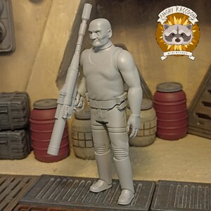 May include: A gray action figure of a bald man in a sleeveless shirt and pants, holding a large rifle. The figure has a belt and knee pads. The background includes barrels and the "Angry Raccoon Miniatures" logo.