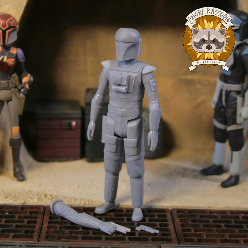 May include: A gray action figure of a character in a helmet and armor, with a detached arm and a small gun on the ground. The figure is standing on a surface with a metal grate. The Angry Raccoon Miniatures logo is visible in the background.