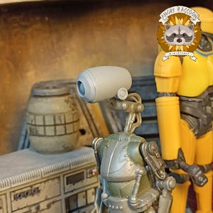 May include: A detailed miniature of a droid with a cylindrical head and a metallic body. The droid is primarily olive green and silver, with a light gray head. A yellow figure stands in the background. The Angry Raccoon Miniatures logo is visible.