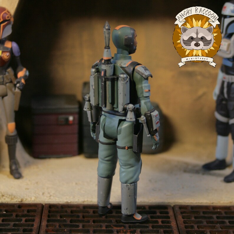 May include: A green and grey action figure of a science fiction character, seen from behind. The figure has a jetpack and other accessories. The Angry Raccoon Miniatures logo is visible in the background.