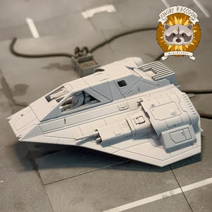 Peut inclure: Maquette de vaisseau spatial miniature gris clair avec des panneaux détaillés, un cockpit et un petit canon. Le modèle est sur une surface grise. Le logo Angry Raccoon Miniatures est dans le coin supérieur droit.