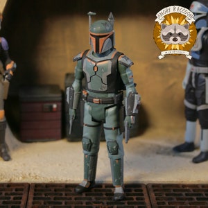 May include: A detailed action figure in green and grey armour with orange detailing. The figure is posed in a diorama, holding a blaster pistol. The figure's helmet has a T-shaped visor. The figure is approximately 15 cm tall.