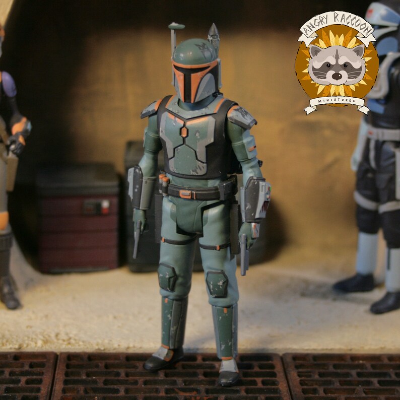 May include: A detailed action figure in green and grey armour with orange detailing. The figure is posed in a diorama, holding a blaster pistol. The figure's helmet has a T-shaped visor. The figure is approximately 15 cm tall.