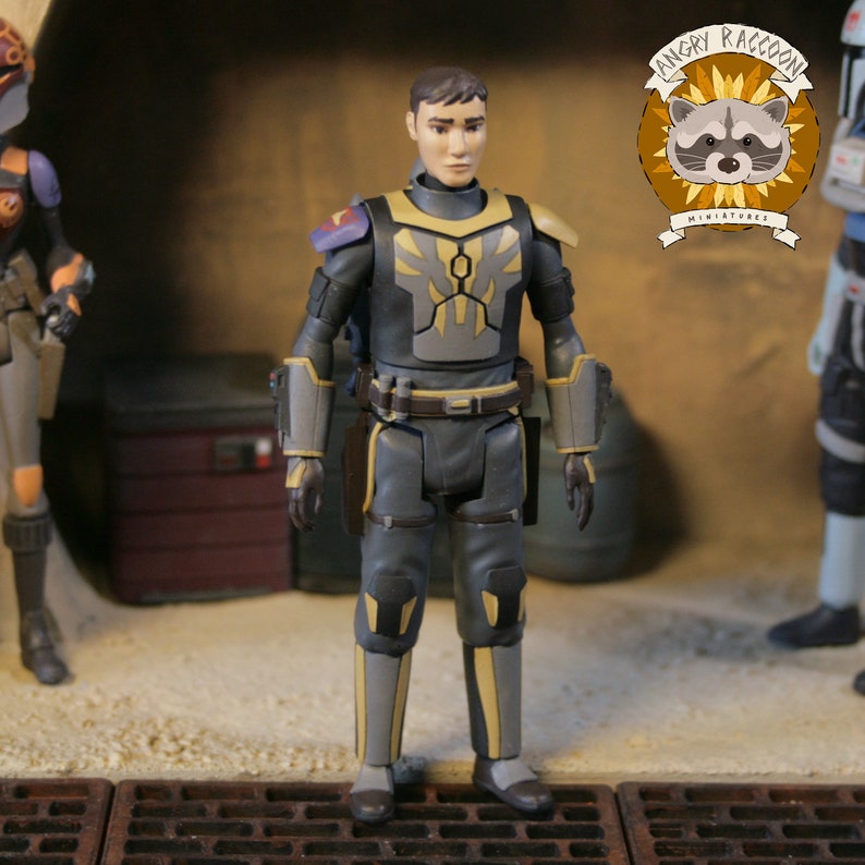 May include: A detailed action figure of a character in a gray and black uniform with yellow accents. The figure has a serious expression and is standing in a diorama setting. The Angry Raccoon Miniatures logo is visible.