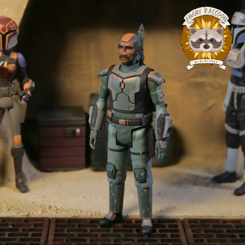 May include: A detailed action figure of a character in a green and black armored suit, with orange detailing. The figure has a helmet with a blade and is standing on a textured surface. The Angry Raccoon Miniatures logo is visible.