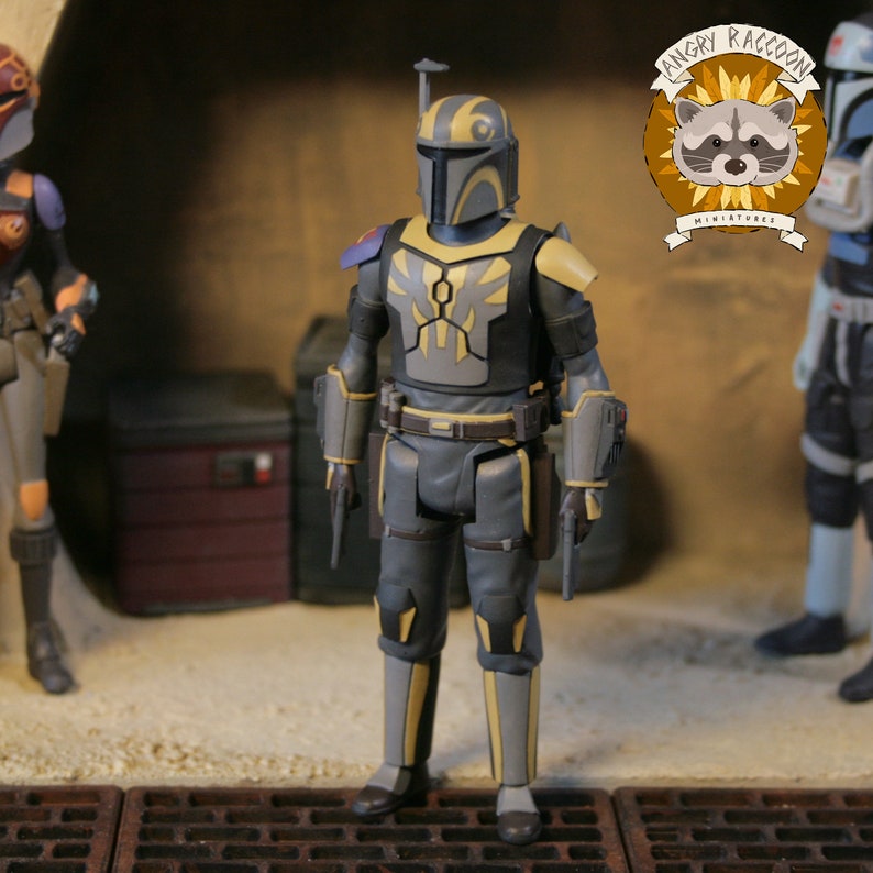 May include: A detailed action figure of a Mandalorian warrior in gray and yellow armor, holding two blasters. The figure has a helmet with a distinctive design and is part of a miniature collection. The Angry Raccoon Miniatures logo is visible.