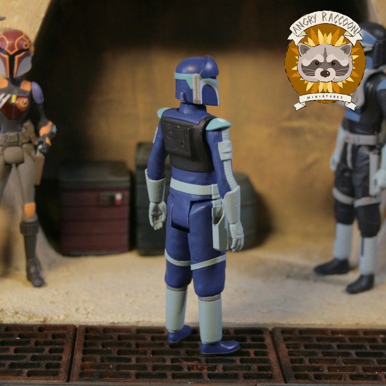 May include: A blue action figure of a character from a science fiction franchise, with light gray accents on the arms and legs. The figure is standing in a diorama setting with other figures and props. The Angry Raccoon Miniatures logo is visible.
