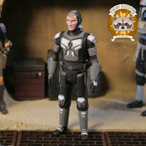 May include: A detailed action figure of a character in a black and gray armored suit. The figure has a blonde hairstyle and is standing in a diorama setting. The Angry Raccoon Miniatures logo is visible in the background.