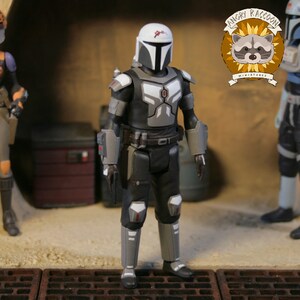 May include: A detailed action figure of a character in black and gray armor with a white helmet and red accents. The figure is standing in a diorama setting with other figures and the "Angry Raccoon Miniatures" logo.