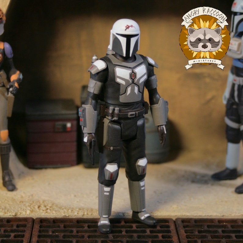 May include: A detailed action figure of a character in black and gray armor with a white helmet and red accents. The figure is standing in a diorama setting with other figures and the "Angry Raccoon Miniatures" logo.
