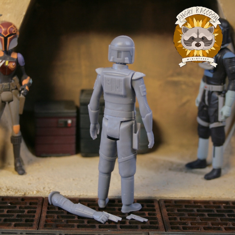 May include: A gray action figure of a character in a helmet and armor, with a detached arm and a small gun on the ground. The figure is standing in front of a brown and tan background, with two other figures in the background. The Angry Raccoon Miniatures logo is in the upper right corner.