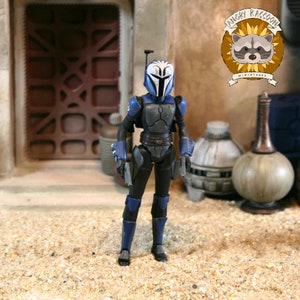 May include: A posable action figure of a warrior wearing blue and black armor with a white helmet featuring blue markings. The figure has a rangefinder on the helmet, dark armor plating on the chest and limbs, and holds two small silver blasters.