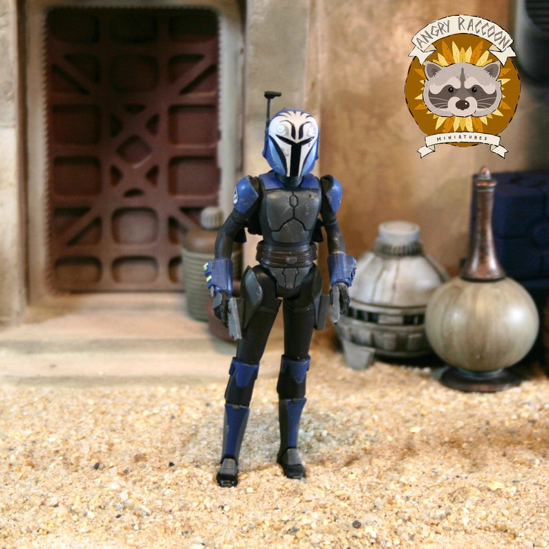 May include: A posable action figure of a warrior wearing blue and black armor with a white helmet featuring blue markings. The figure has a rangefinder on the helmet, dark armor plating on the chest and limbs, and holds two small silver blasters.