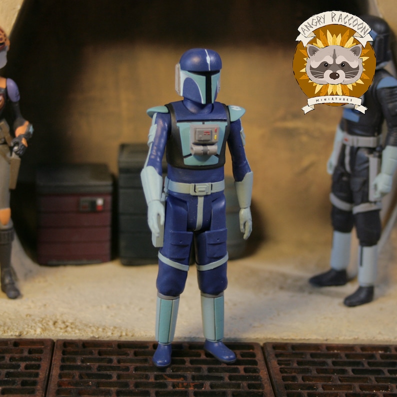May include: A blue action figure with light blue accents, part of a series of miniature figures. The figure is wearing a helmet and armor, standing in a detailed diorama setting. The Angry Raccoon Miniatures logo is visible.