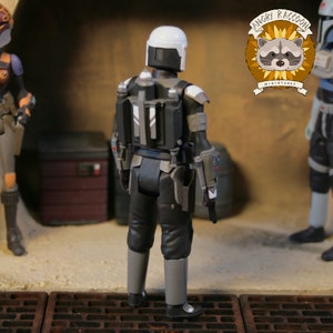 May include: A black and gray action figure of a character from a science fiction series, viewed from the back. The figure wears a white helmet, gray boots, and a black backpack. The figure is standing on a textured surface with other figures in the background.