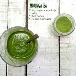 May include: A glass of green moringa tea with a spoon in it, next to a jar of moringa powder. The text on the image reads "MORINGA TEA * 1 tsp organic moringa powder * 1 tsp honey * 1/2 cup hot water"