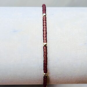 May include: A beaded bracelet featuring small, dark red beads strung together with gold-colored accents. The bracelet is displayed vertically against a light gray background, showcasing its simple yet elegant design. The beads are evenly spaced, creating a symmetrical pattern.