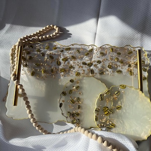May include: A set of three white resin serving trays with gold accents and a scattered pattern of gold flakes and coffee beans. The trays are on a white surface with a wooden bead necklace.