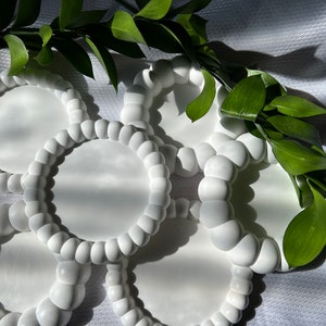 May include: Five white ceramic coasters with a raised, beaded edge. The coasters are arranged in a circle on a white surface.