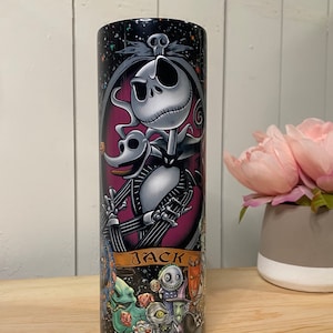 Peut inclure: Un gobelet noir avec une illustration d&eacute;taill&eacute;e de Jack Skellington de L'&Eacute;trange No&euml;l de Monsieur Jack. Le design comprend Jack, Z&eacute;ro et d'autres personnages, avec le nom "JACK". Le gobelet est cylindrique et pos&eacute; sur une surface en bois.