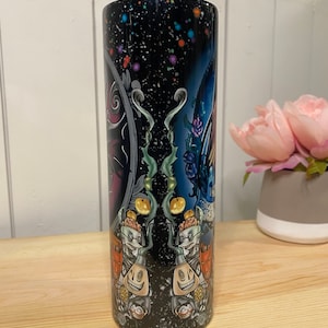 Peut inclure: Un gobelet cylindrique avec un fond bleu fonc&eacute; parsem&eacute; de points color&eacute;s. Le gobelet pr&eacute;sente un motif effrayant avec des personnages, des citrouilles et des &eacute;l&eacute;ments floraux. Les mots "There's" sont visibles sur le motif.