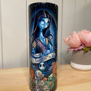 Peut inclure: Un gobelet cylindrique repr&eacute;sentant un personnage &agrave; la peau bleue avec de longs cheveux noirs, tenant une fleur sombre. Le gobelet a un fond bleu fonc&eacute; avec le texte "I sense there's something in the wind." Le bas du gobelet pr&eacute;sente d'autres personnages.