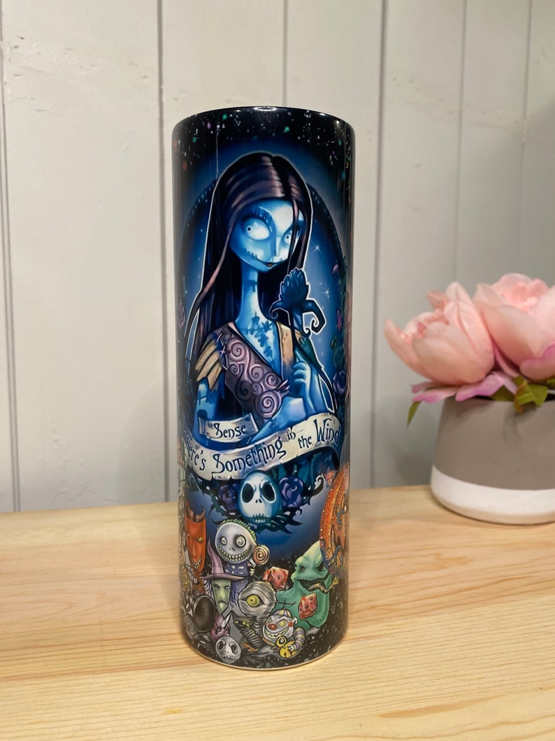 Peut inclure: Un gobelet cylindrique repr&eacute;sentant un personnage &agrave; la peau bleue avec de longs cheveux noirs, tenant une fleur sombre. Le gobelet a un fond bleu fonc&eacute; avec le texte "I sense there's something in the wind." Le bas du gobelet pr&eacute;sente d'autres personnages.