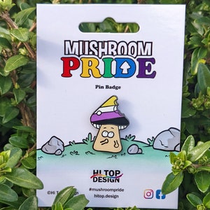 Pode incluir: Um broche de segurança com um cogumelo de desenho animado com uma bandeira de orgulho genderqueer amarela, roxa e branca em seu chapéu. O texto "Mushroom Pride" é impresso em cores do arco-íris acima do cogumelo. O texto "Pin Badge" é impresso abaixo do cogumelo. O texto "Hi Top Design" é impresso abaixo do cogumelo com a hashtag "#mushroompride" e o site "hitop.design".