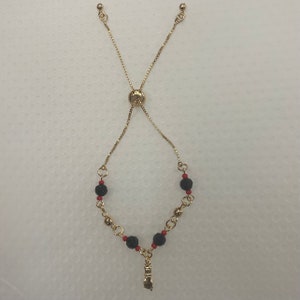May include: A gold chain bracelet with black and red beads and a gold charm in the shape of a hand.